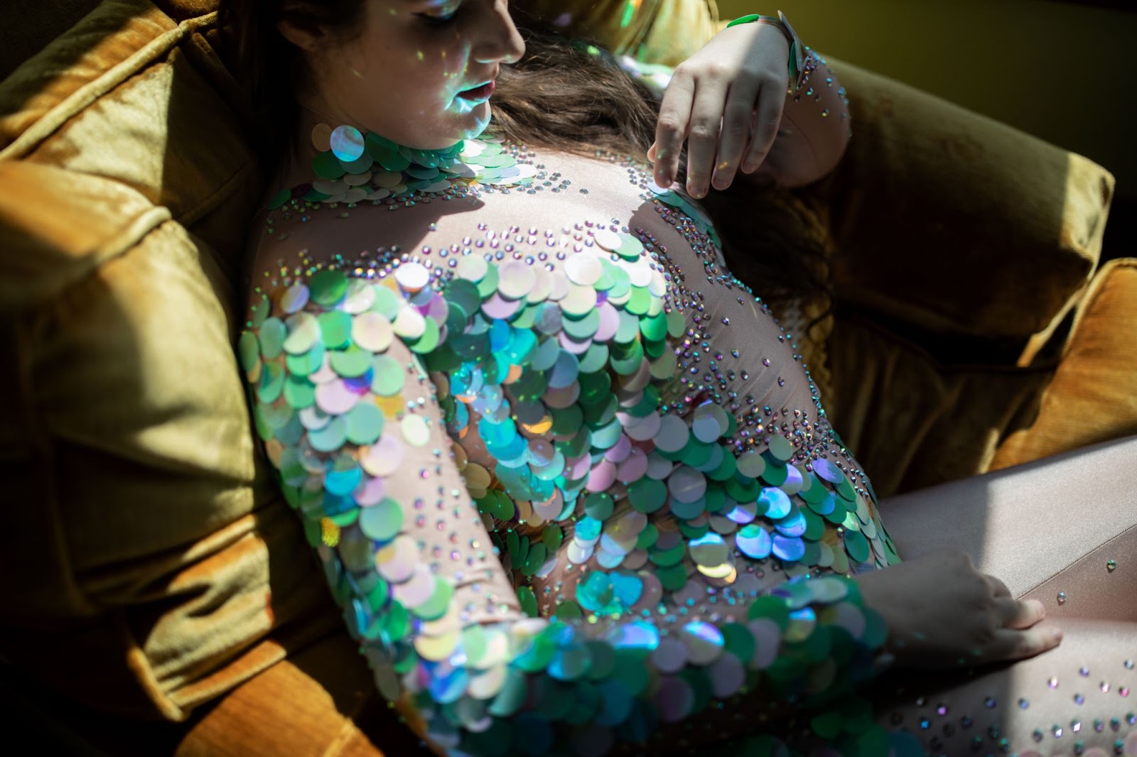 Image: A portrait of performer Secret Mermaid. They are pictured sitting on a gold-toned velvet couch, wearing an iridescent fish scale-like sequined and bejeweled costume, crafted by fellow burlesque performer and cabaret star, UK's Talulah Blue, 2023. Photo by Erica McKeehen.