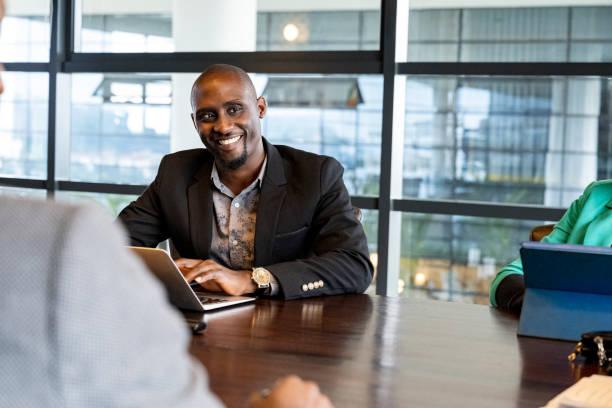 Confident Black businessman in board room meeting Mid adult executive sitting at conference table with colleagues, pausing from typing on laptop to smile at off-camera associate. kenyan businessman stock pictures, royalty-free photos & images