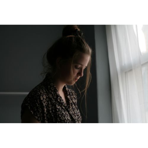 A sad looking young woman standing next to a window with her head down.
