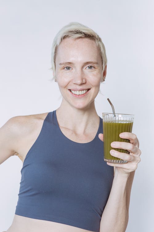 Free Positive young female with dyed short hair wearing activewear standing with hand on waist with glass of green smoothie Stock Photo