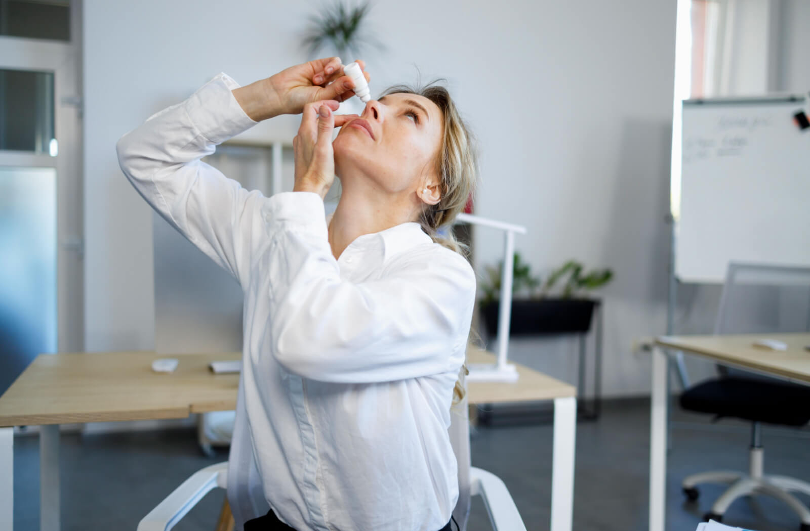 A woman applying eye drops to her eyes while sitting in her office.