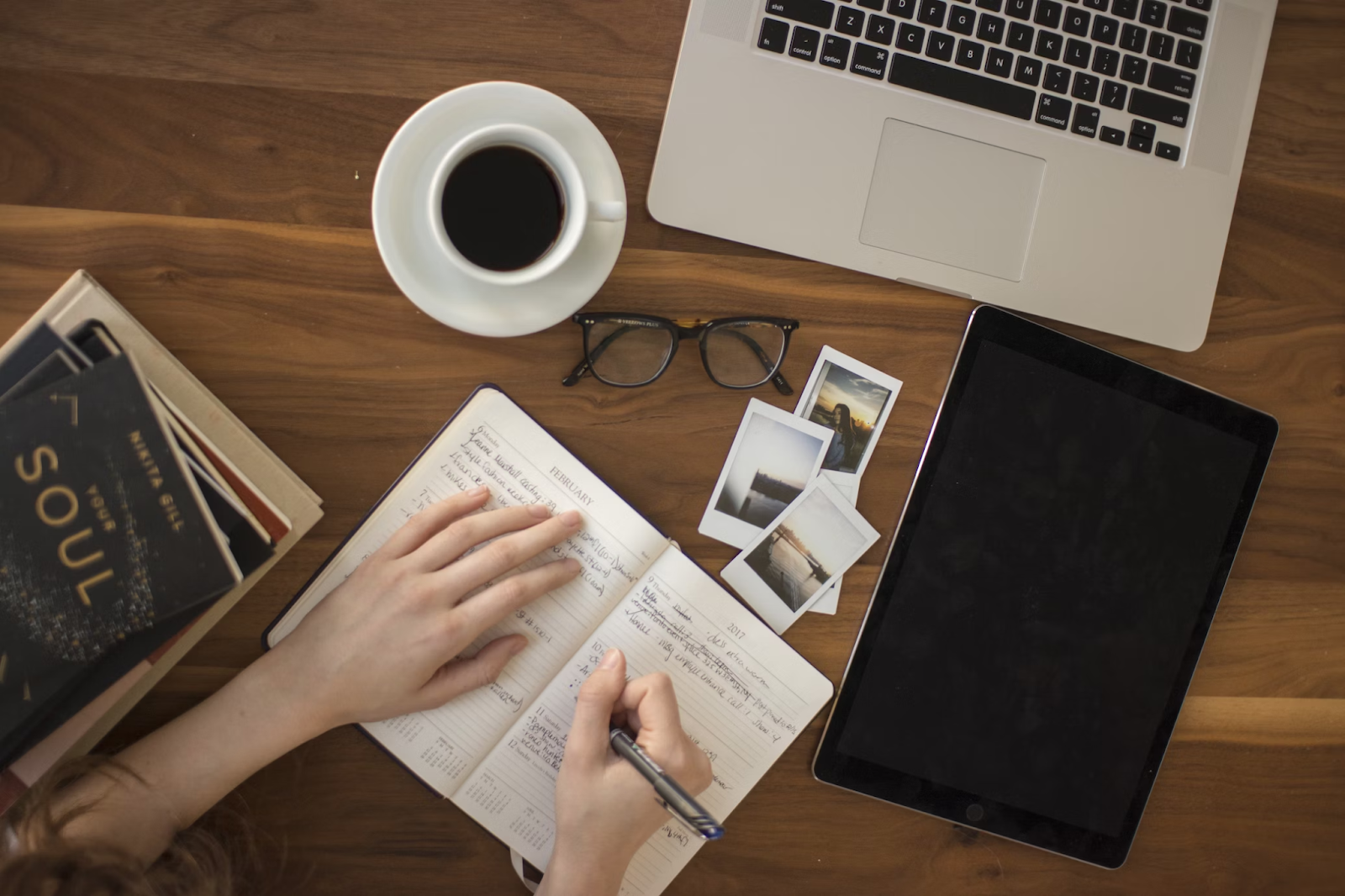 Mesa de madeira com um laptop, um tablet, livros, uma xícara de café e um caderno. Uma pessoa está sentada na mesa escrevendo no caderno