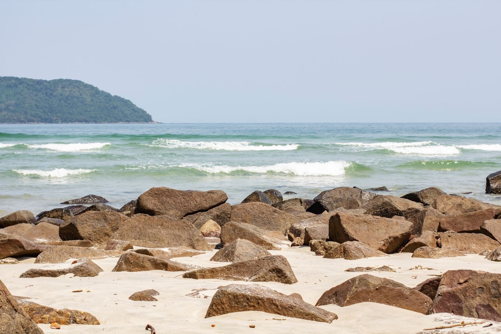 Mar da praia de Juquehy. A água de tonalidade azul clara forma pequenas ondas que quebram em um trecho de areia branca com muitas pedras escuras.