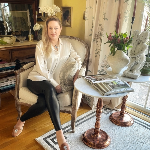 Leah Kroeber sits on a French style bergere in her living room next to two on pedestal side tables with books and decorative items such as vase and sculpture upon them.