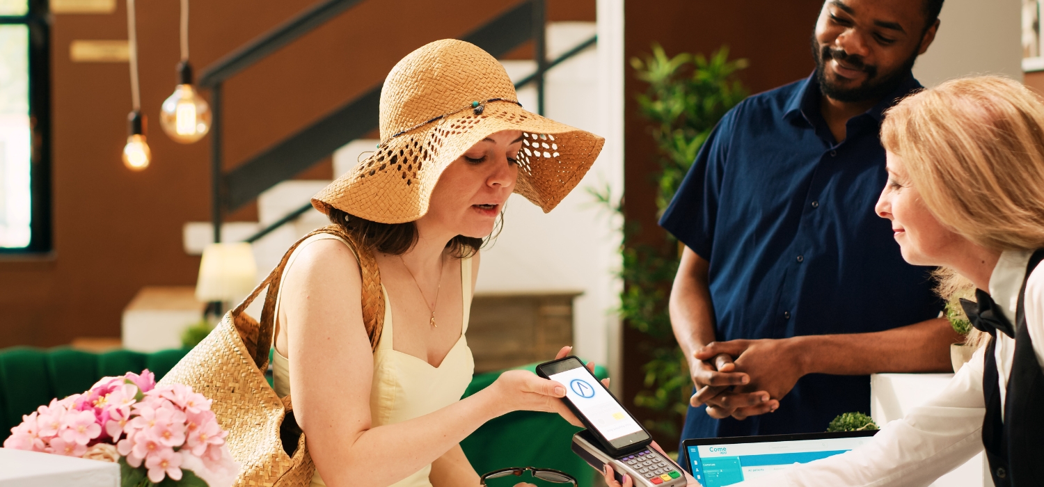 A woman wearing a summer flower hat and yellow dress is using NFC technology in 2023 from the GOTAP smart tag on the back of her smartphone to a waiter at a luxury restaurant in Bali.