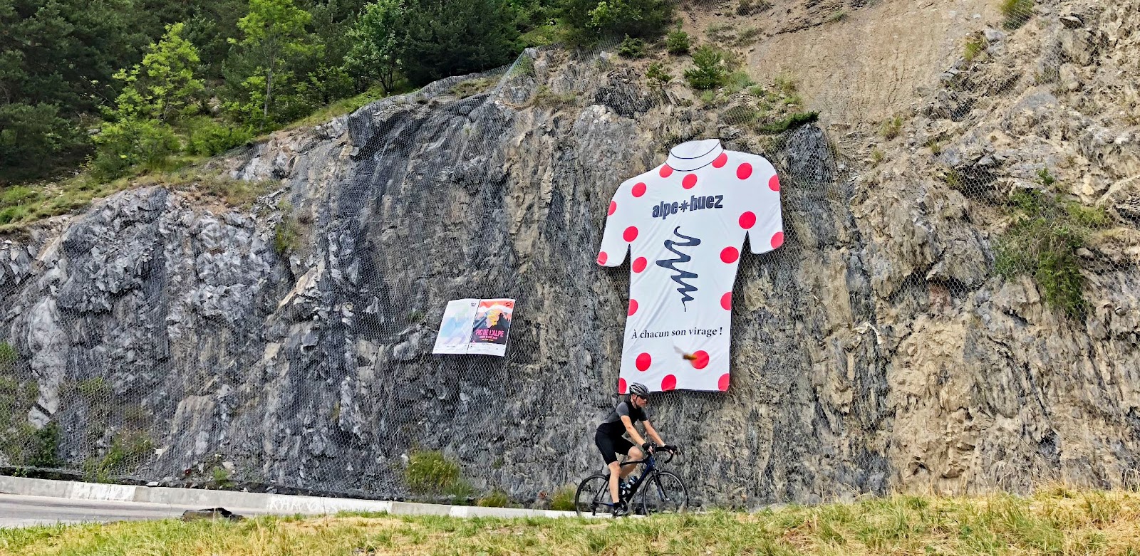 cyclist rides by large polka dot jersey sign on rock wall, Alpe d'Huez
