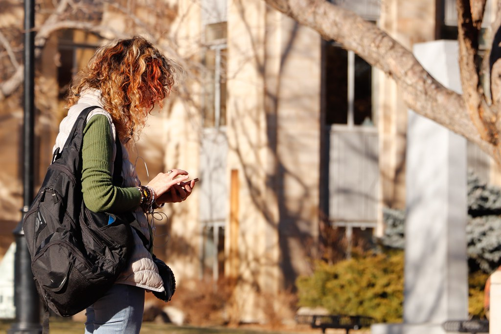 A woman wears a black backpack and looks down at her phone - Benefits of Going To Community College