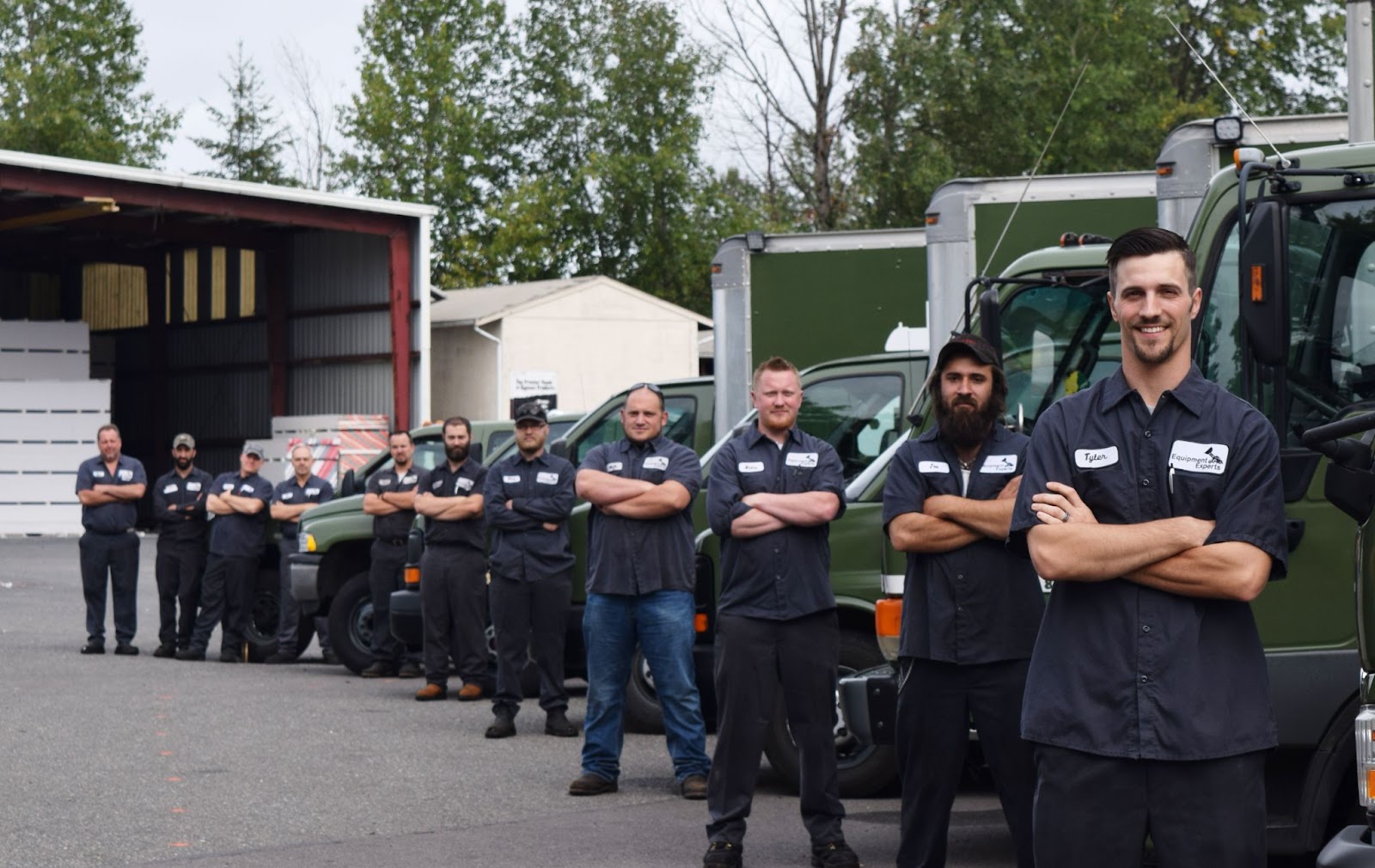 A team of diesel repair technicians with their arms crossed in a diagonal line while standing next to semi-trucks