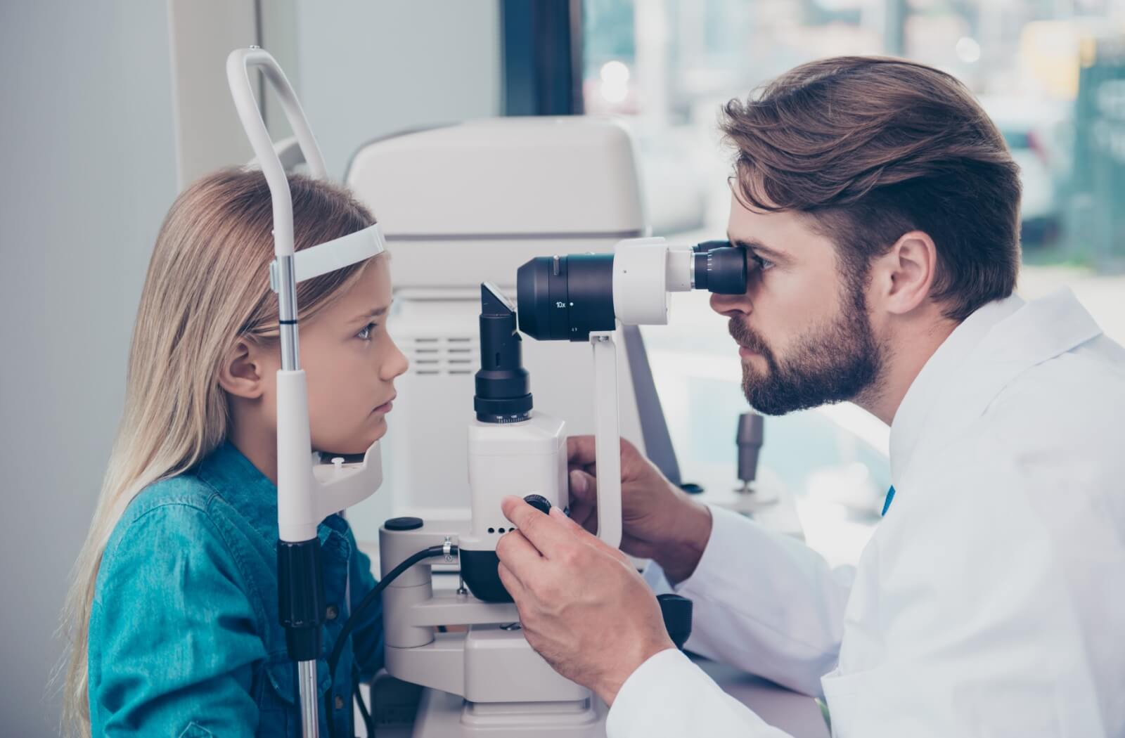 A male optometrist uses a medical device to examine the eyes of a young child and look for potential eye problems.