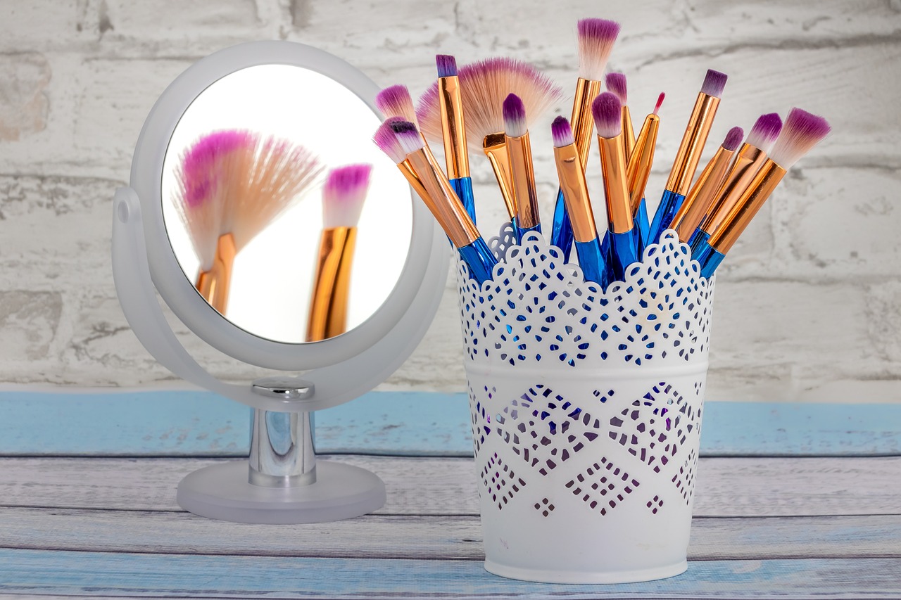 Round mirror and a white container of blue, gold and pink makeup brushes on a blue and white surface