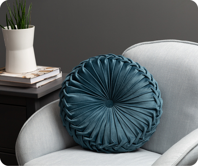 Our dark teal Pin-Tuck Round Corduroy Cushion in Ocean shown styled on an armchair.