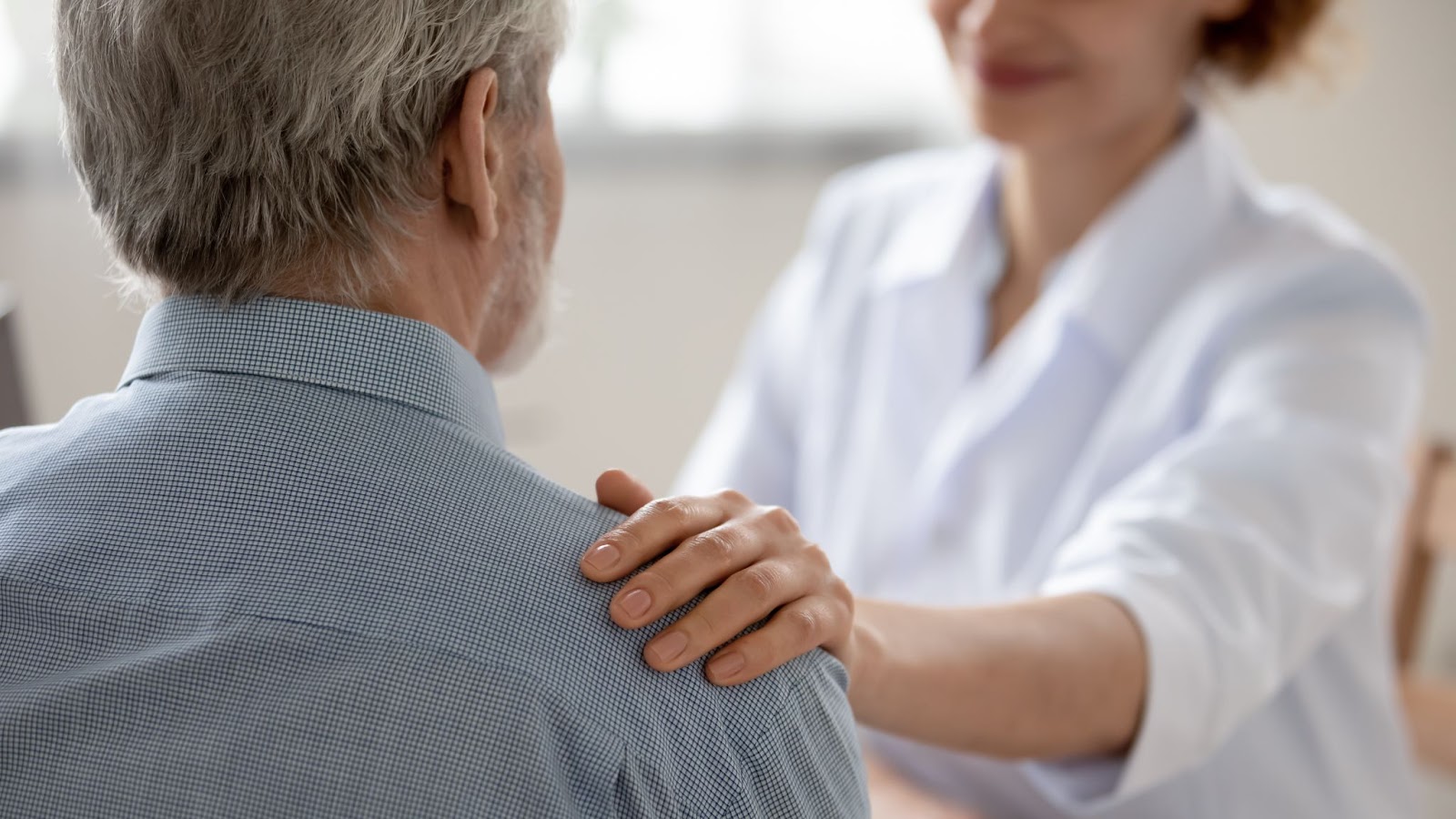 An adult child offers a supportive smile while placing her hand on the shoulder of her older adult father.