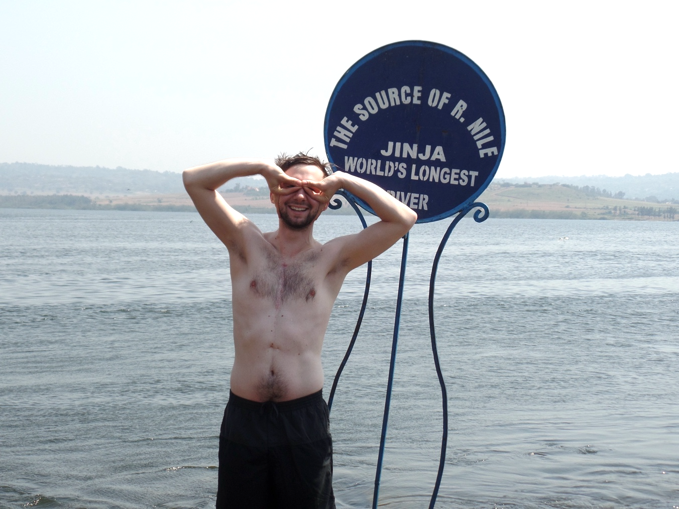 Stephan poses, making a goofy glasses shape with his fingers, in front of a sign reading "The source of R. Nile, Jinja, World's Longest River." The river flows behind him.