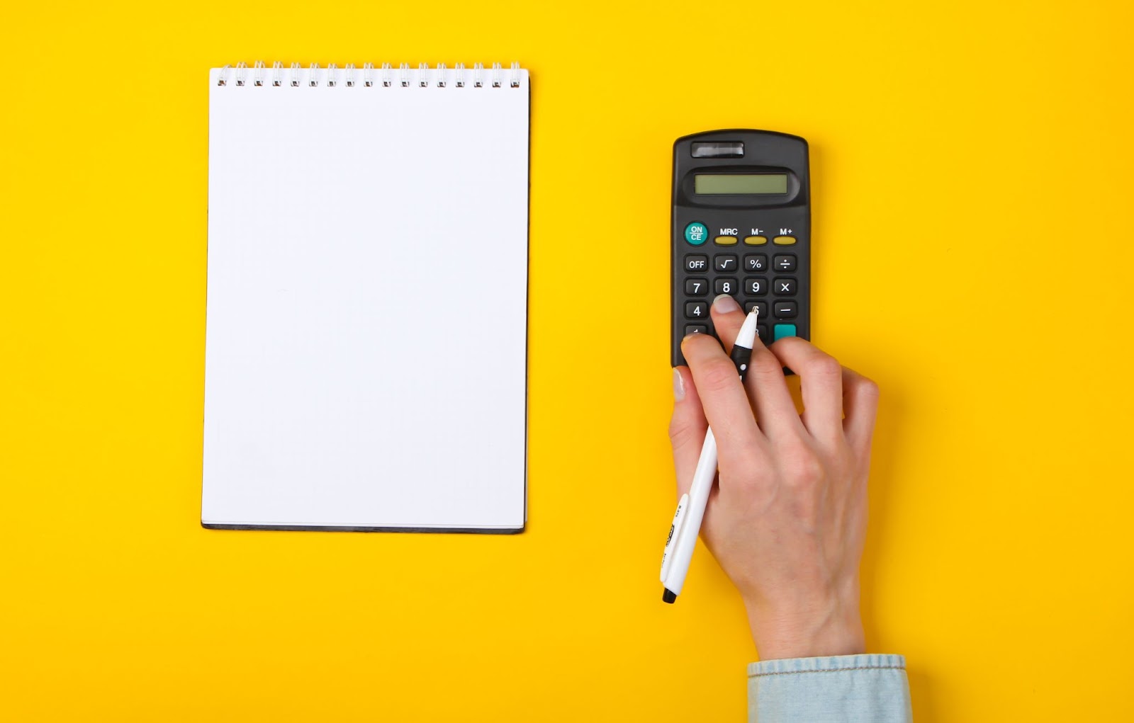 Imagem focada em uma mão que está mexendo em uma calculadora, enquanto um caderno com folha em branco está ao lado do objeto. A mão é branca e há uma caneta apoiada entre os dedos. A superfície onde tudo está apoiado é amarela.