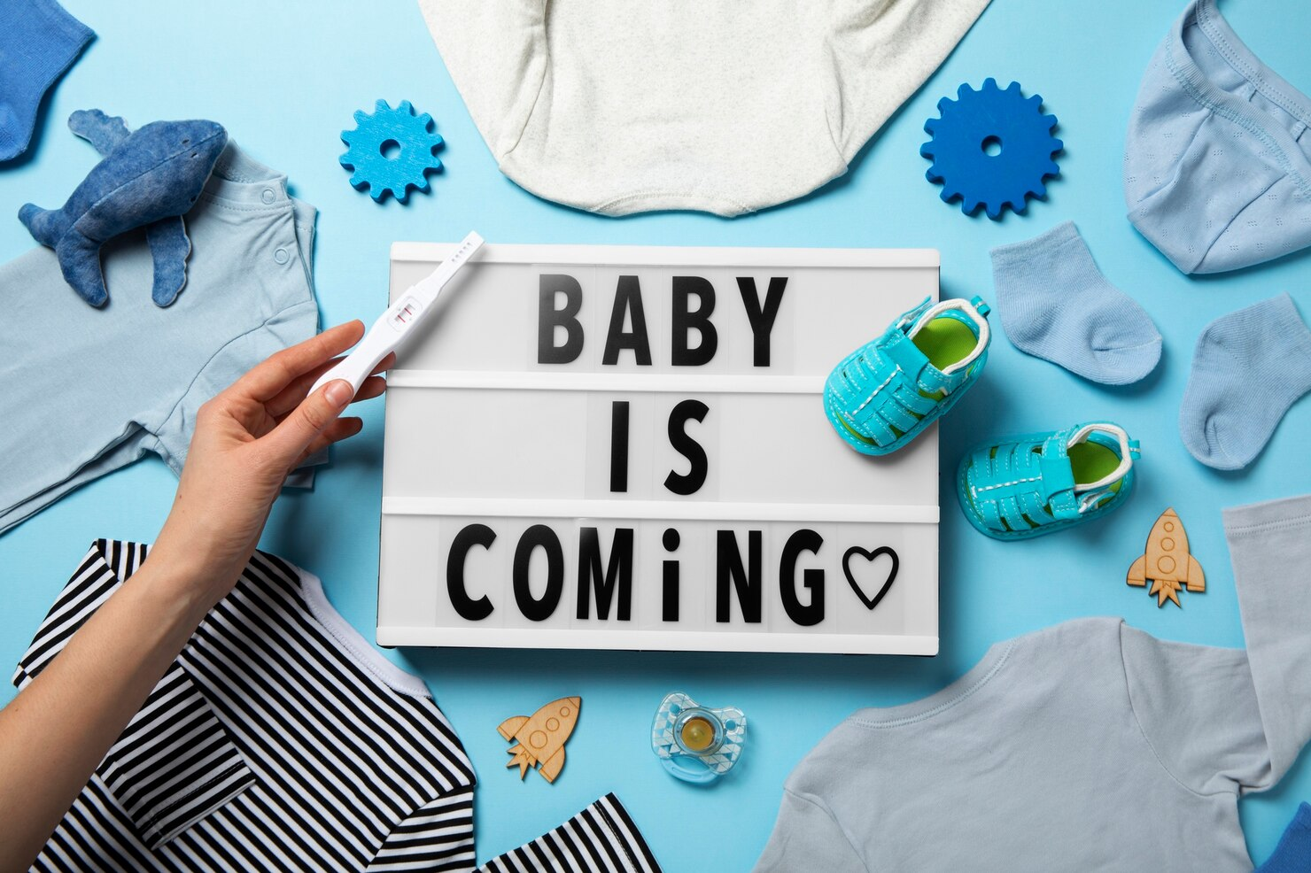 A woman holding a positive pregnancy test against a sign that says "baby is coming" with baby things around.