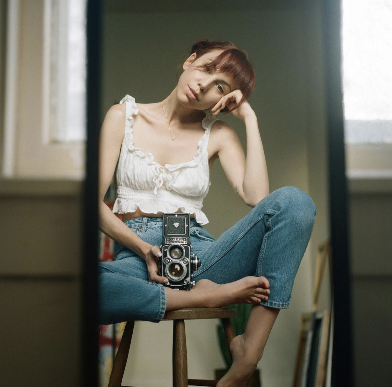 A self portrait taken by photographer Allister Ann, sitting on a wood stool holding a vintage camera, resting her head on her hand.