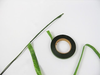 On a white paper tabletop, a floral wire lays partially wrapped with corsage tape. The roll of corsage tape lays in the middle of the picture as its unrolled tape is weaved around the area. 