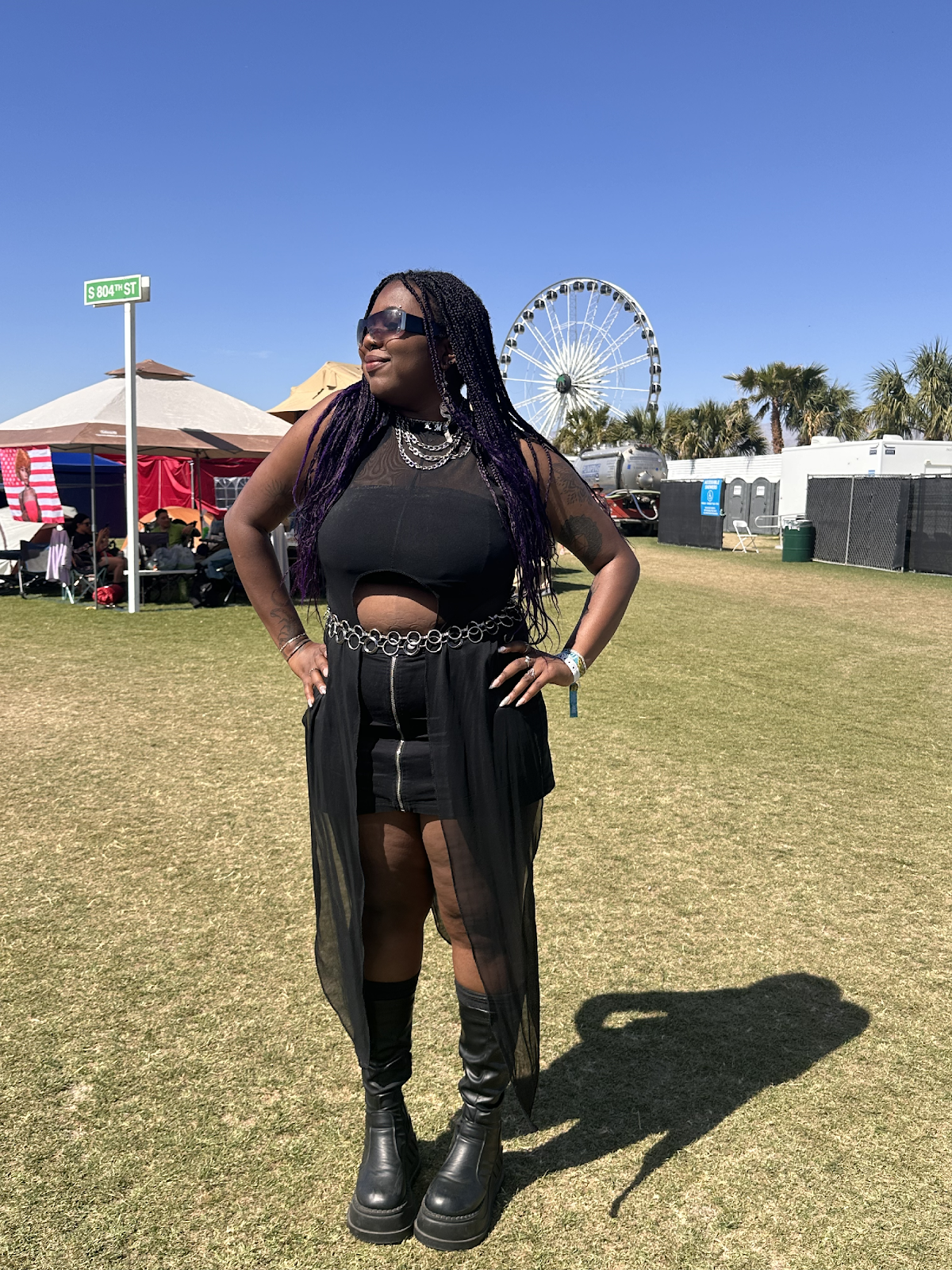Cameryn, a dark-skinned woman participating in the Accessible+ program, is standing with her hands on her hips in a grassy field at Coachella. She has long black and purple locs, is wearing black shades, a black top with a circle cutout on the bottom, an intricate belt, a black zip skirt, a black sheer long skirt, and black boots with silver necklaces. The sun is bright, casting a hard shadow of her onto the ground.