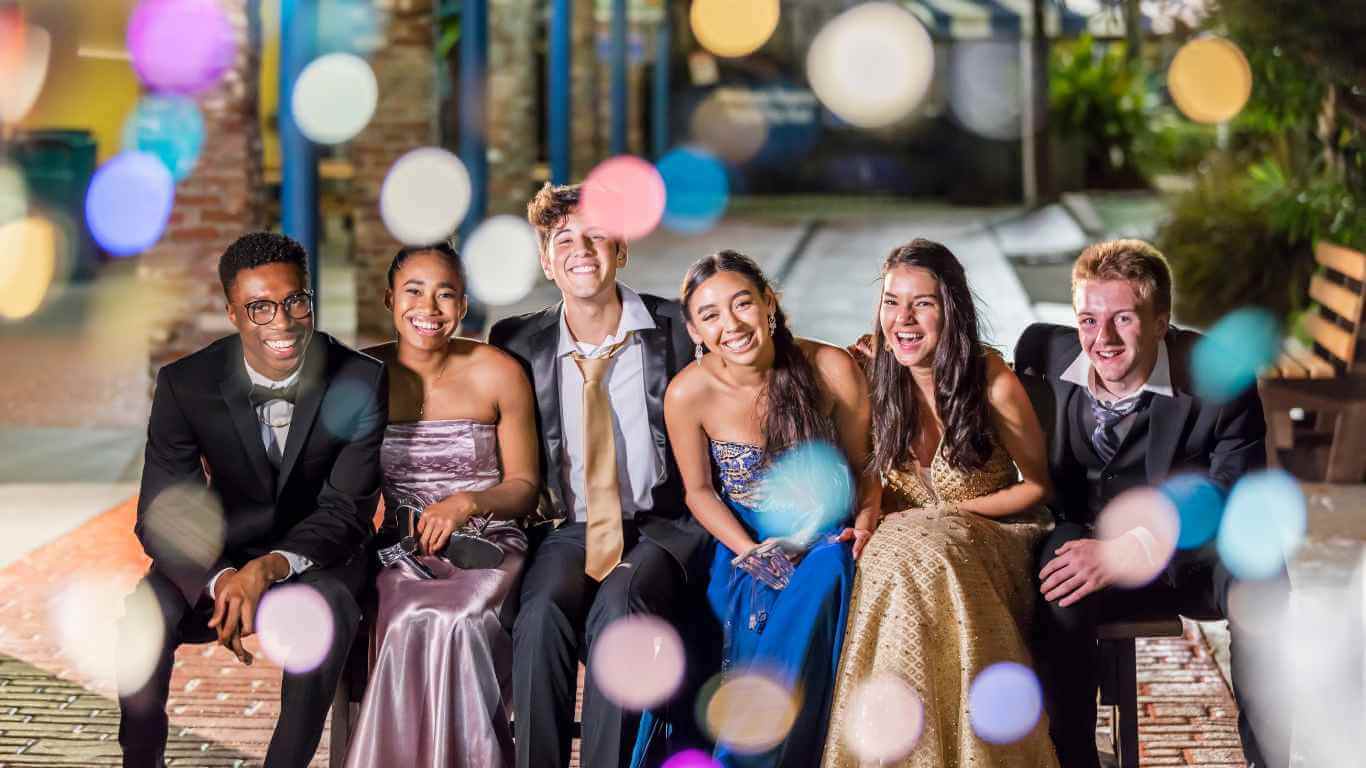 Prom Captions For Instagram - A group of five happy teenagers in formal wear, laughing and sitting together at prom night, with colorful bokeh lights in the background.
