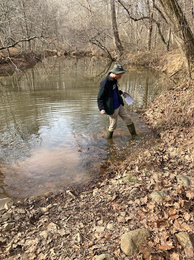 Beautiful blonde young woman fishing near creek wearing coveralls -  standing next to tree Stock Photo