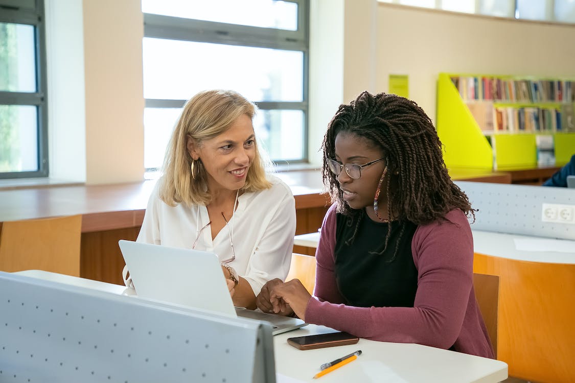 Free Positive adult female teacher explaining task to young black student with Afro braids doing assignment on laptop in modern classroom Stock Photo