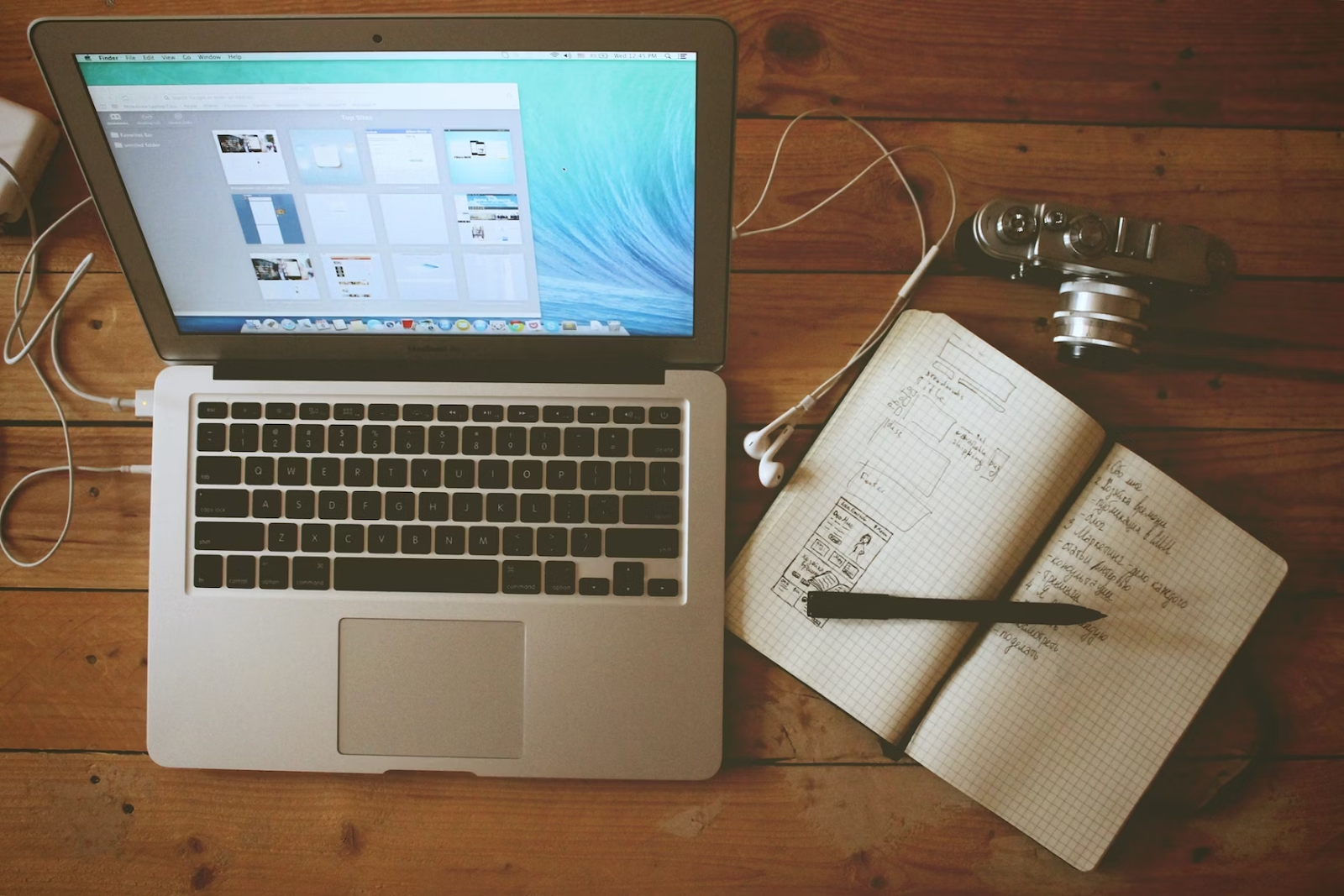 an overhead shot of a laptop with design software on a brown table with a notebook, pen, camera, and headphones