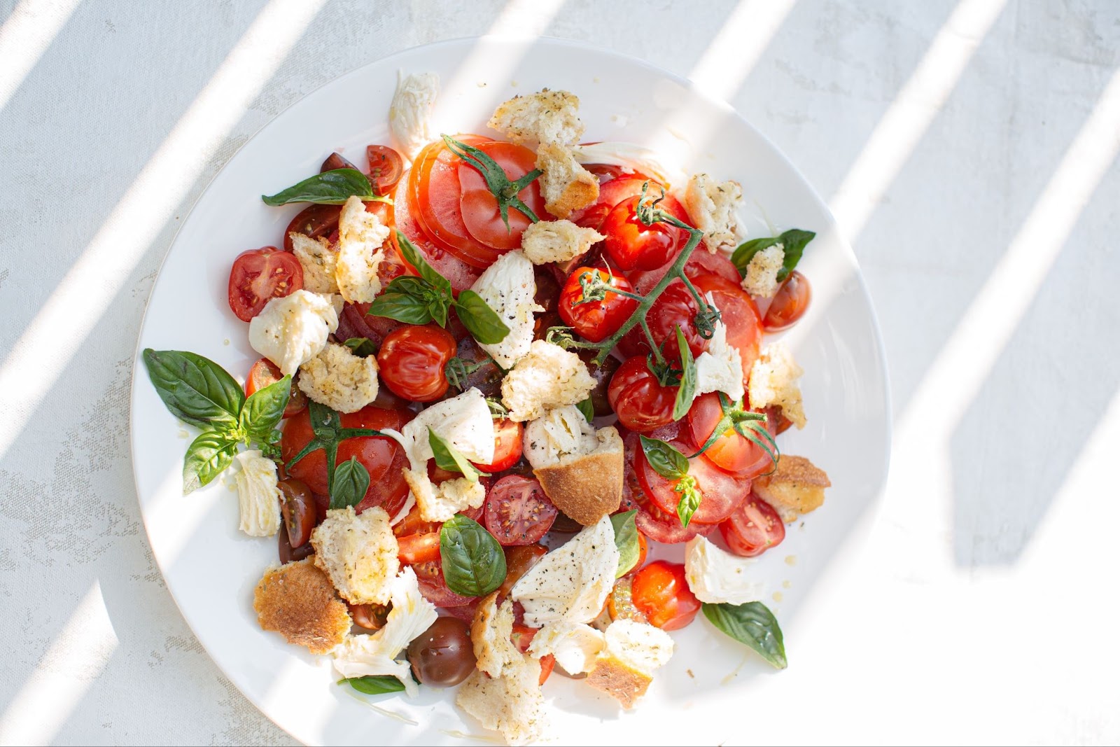 a top-down view of an Italian caprese salad, with mozzarella, croutons, tomatoes, and basil, all in a white plate on a white marble countertop