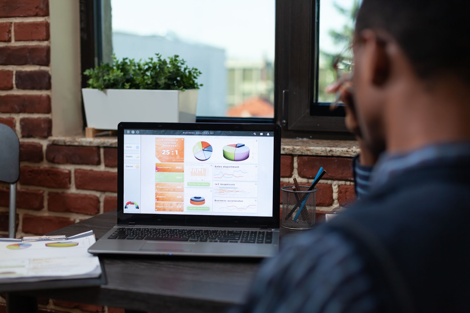 A laptop screen showing sales results charts positioned on a desk near a glass window while a male executive, with his back to the camera, analyzes the data on the screen; a plant box decorates the windowsill.