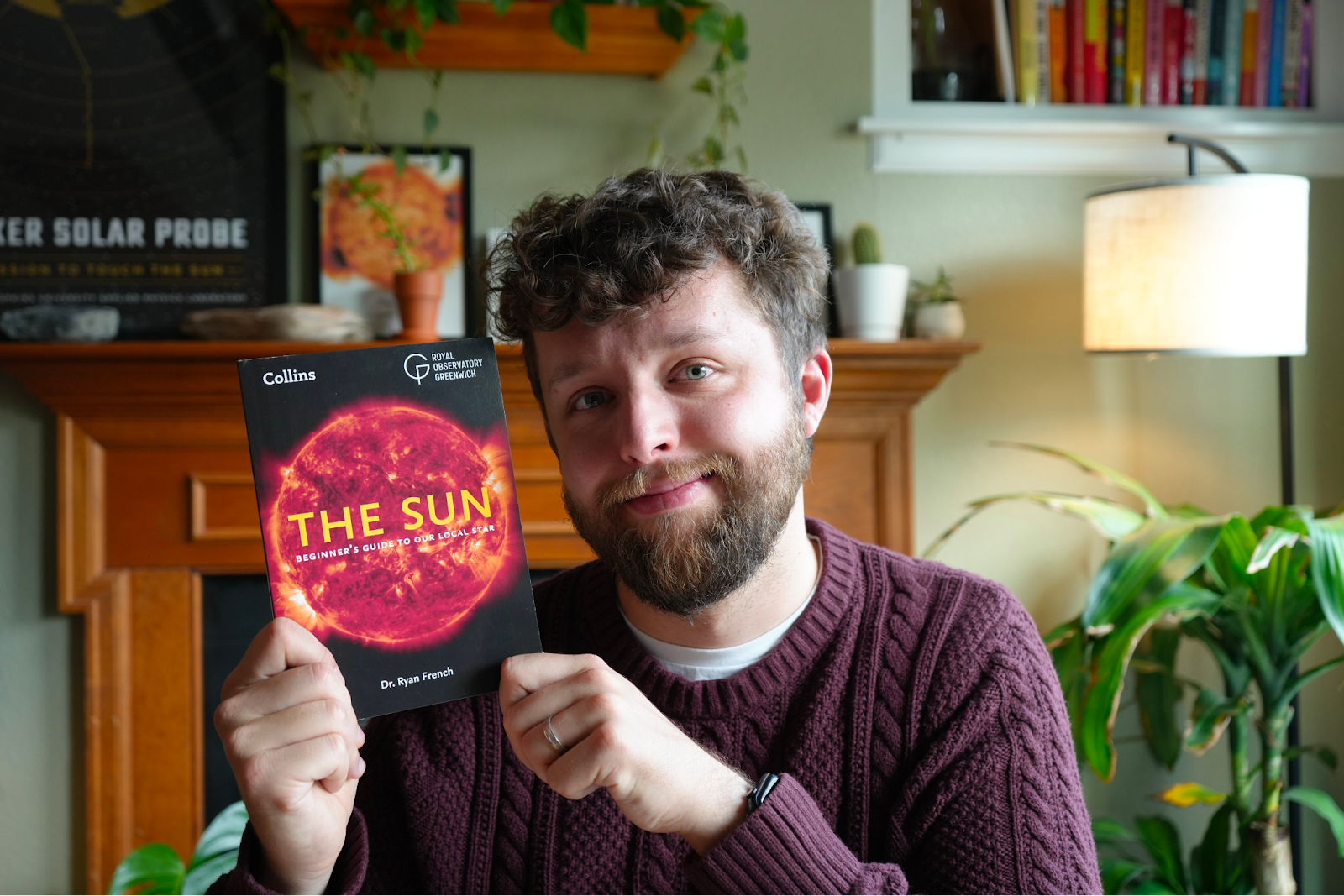 A man with brown curly hair and a beard wearing a burgundy cable knit sweater, holding up a copy of the book The Sun. The book is black with a bright orange and red image of the sun, and "the sun" in all caps yellow text.