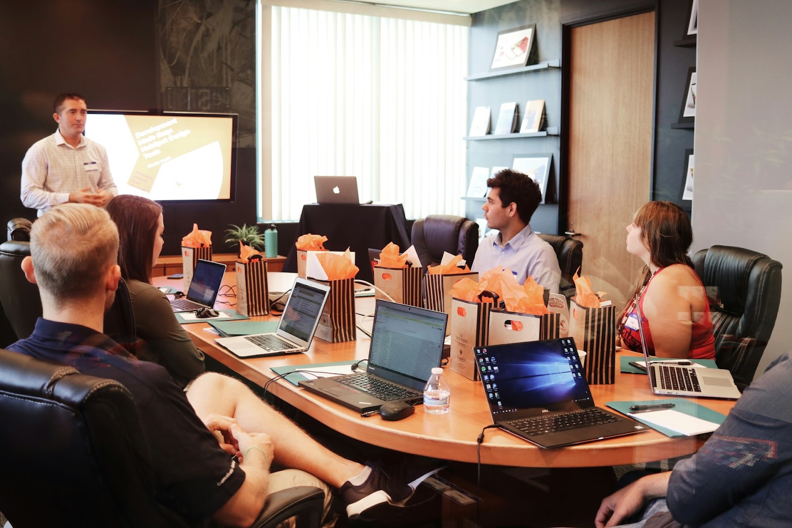 Professionals at a conference table with laptops, watching a presentation given by a person standing in front.