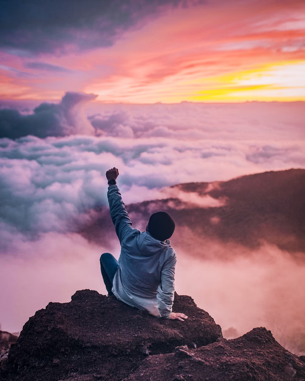 A Boy Sitting on a Mountain in Colorful Clouds With His Fist In The Air