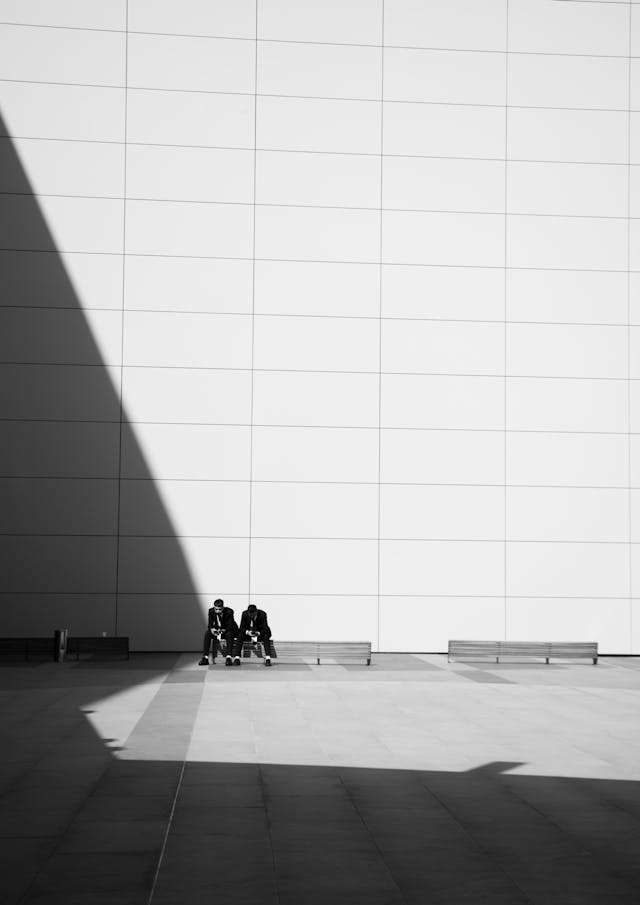 A black and white photograph of two men sitting on a bench in front of a tall austere building designed using parametric design principles