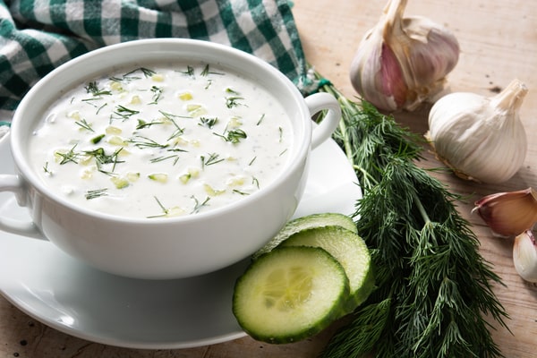 A bowl of alfredo sauce with cucumber, herbs, onions and garlic on the side