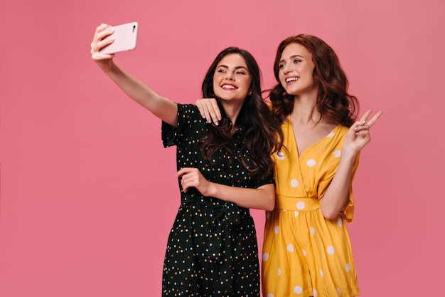 Free photo curly ladies in stylish summer dresses take selfie on pink background beautiful girlfriends in bright overalls posing for photo