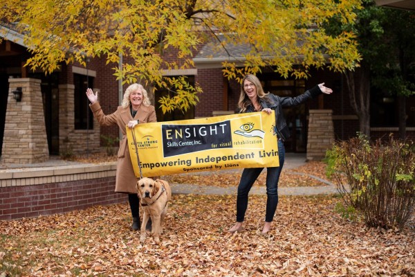 Two women standing in front of a brick and concrete building with an “Ensight Skills Center” yellow banner and a golden retriever on a leash