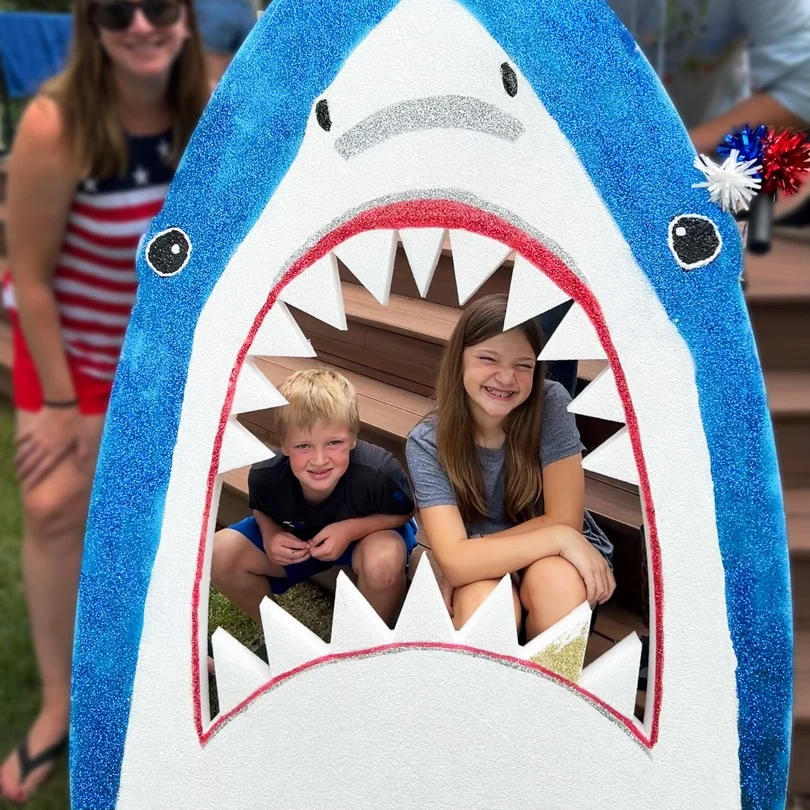 a foam cut-out of a shark. inside the open mouth, two children pose for a photo.