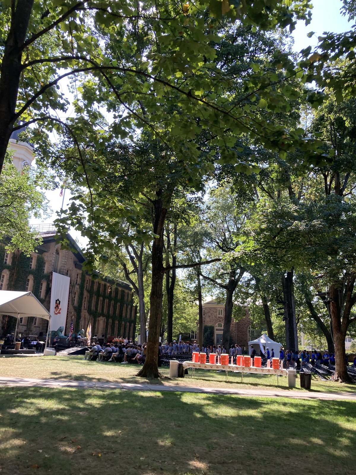 Large audience is seated in chairs set up under trees in front of pre-revolutionary building (Nassau Hall)