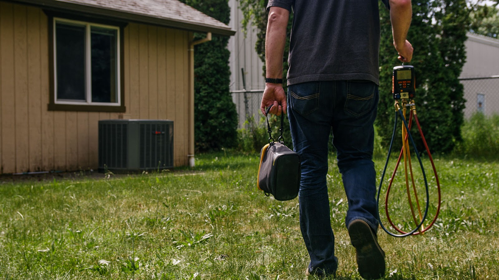An HVAC tech walks towards a house with tools in-hand, prepared to start working.