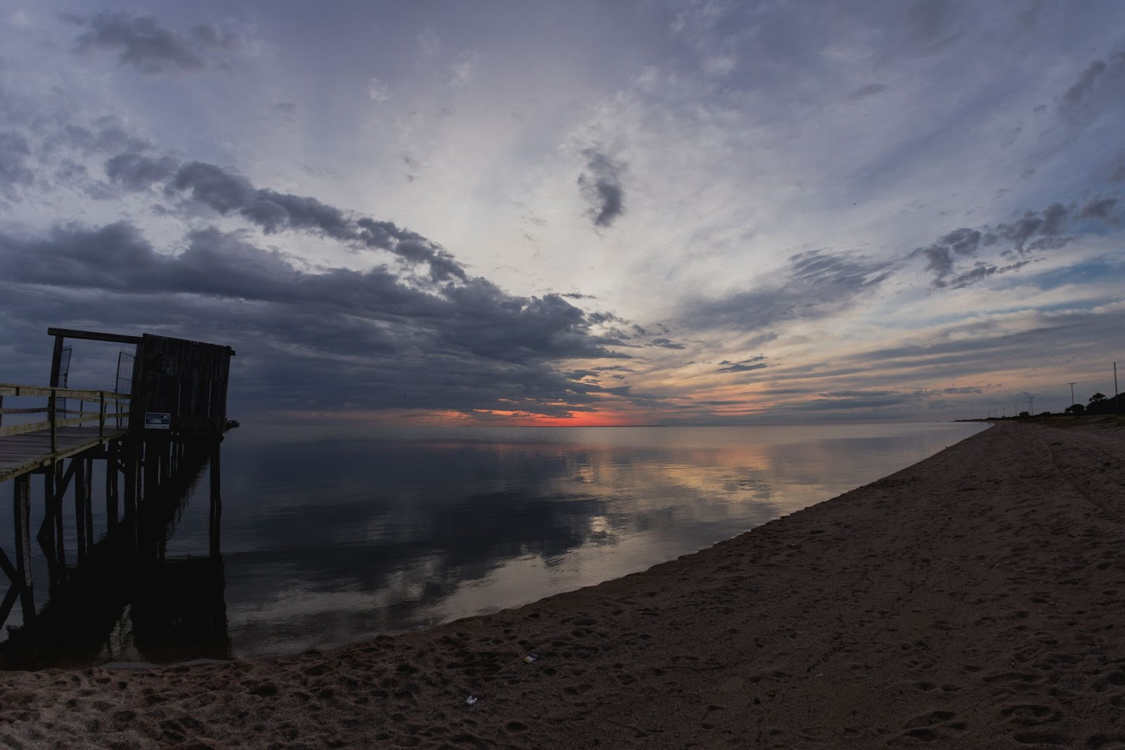 Praia de lago ao entardecer, com nuvens cobrindo o sol, aparecendo apenas a iluminação alaranjada ao horizonte.