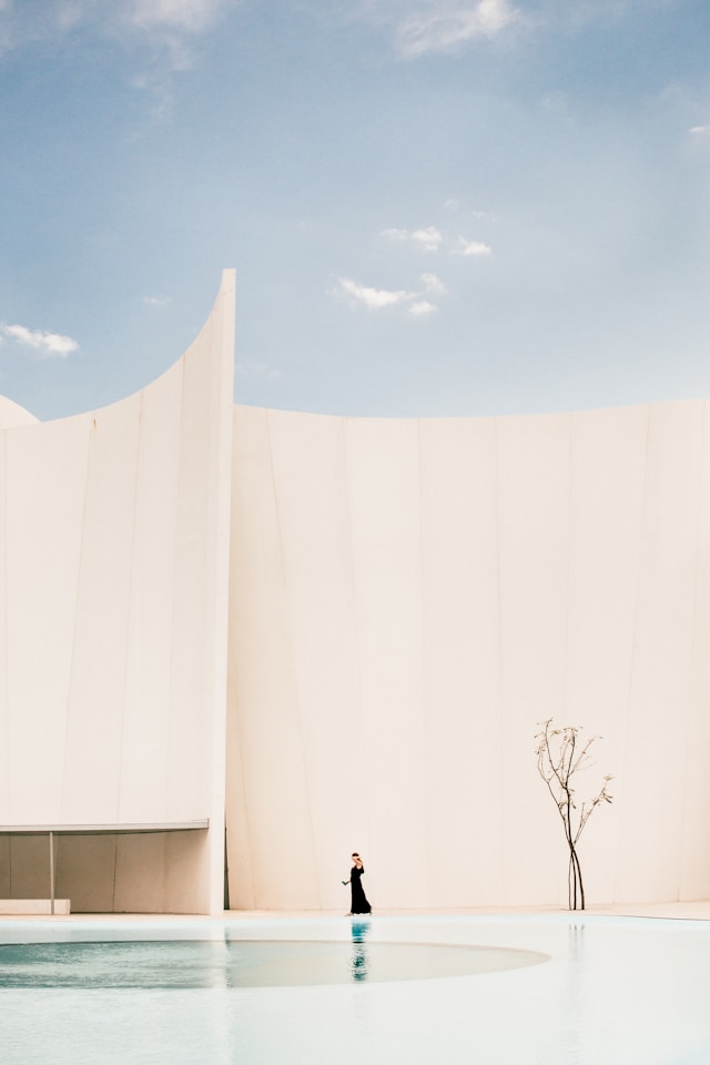 An off white curved building with a silhouette of a woman and a tree in front of it there is a water body in front of the building designed using parametric design principles
