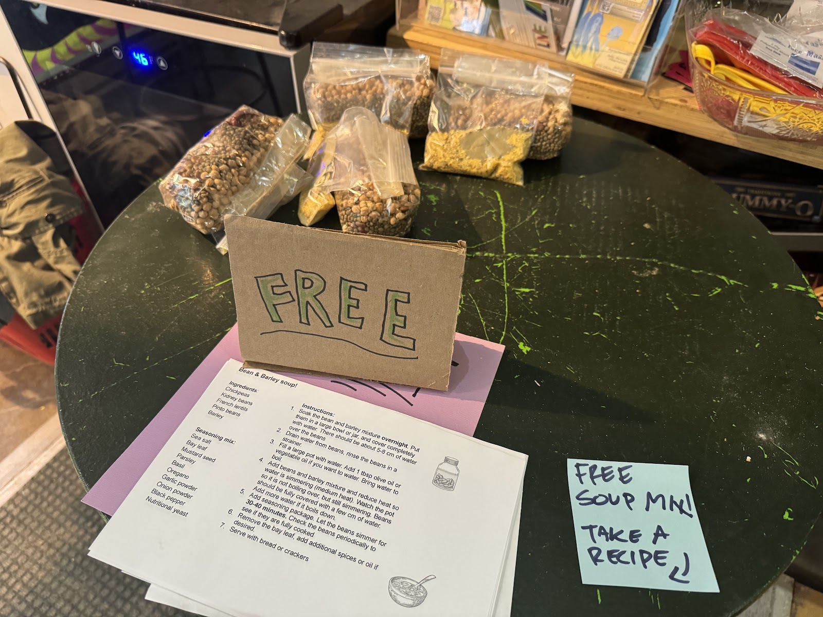 A table with bags of beans and soup ingredients, a handmade sign that says “FREE” and “Bean & Barley soup” recipes. A post-it note says “FREE SOUP MIX! TAKE A RECIPE.”