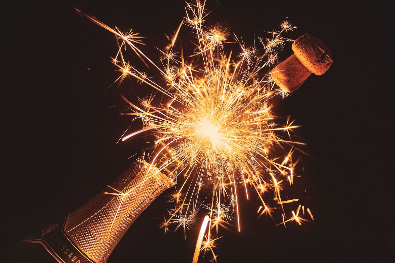 A bottle of champagne opening with sparkles coming out and the cork shooting off on a black background.