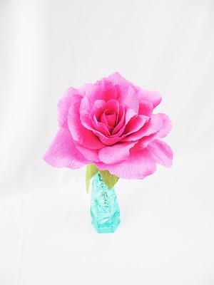A blossoming pink rose, with paper leaves, is in a small turquoise vase on a white background.