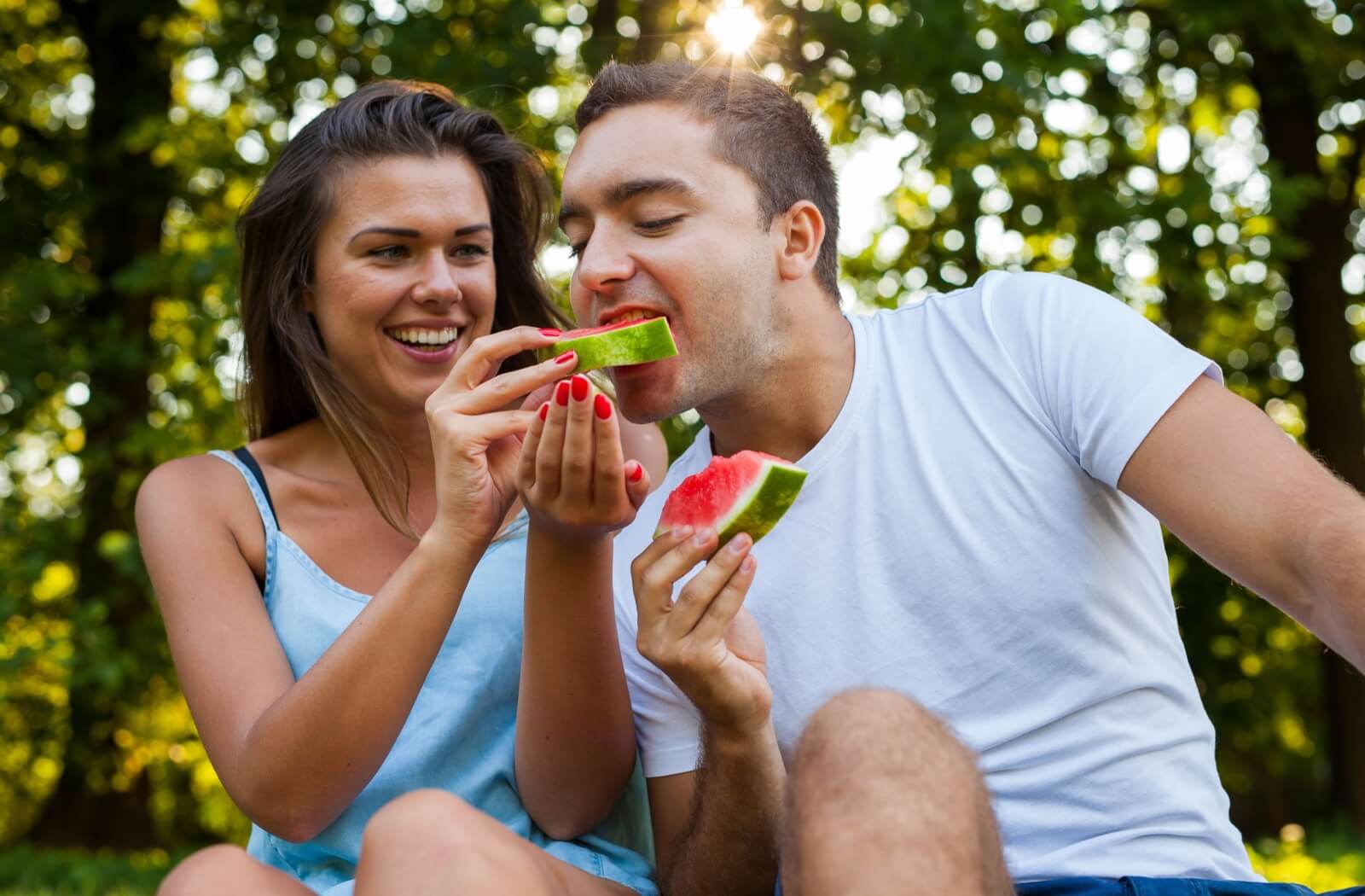 A young man taking a bite from a watermelon held by a woman beside him.