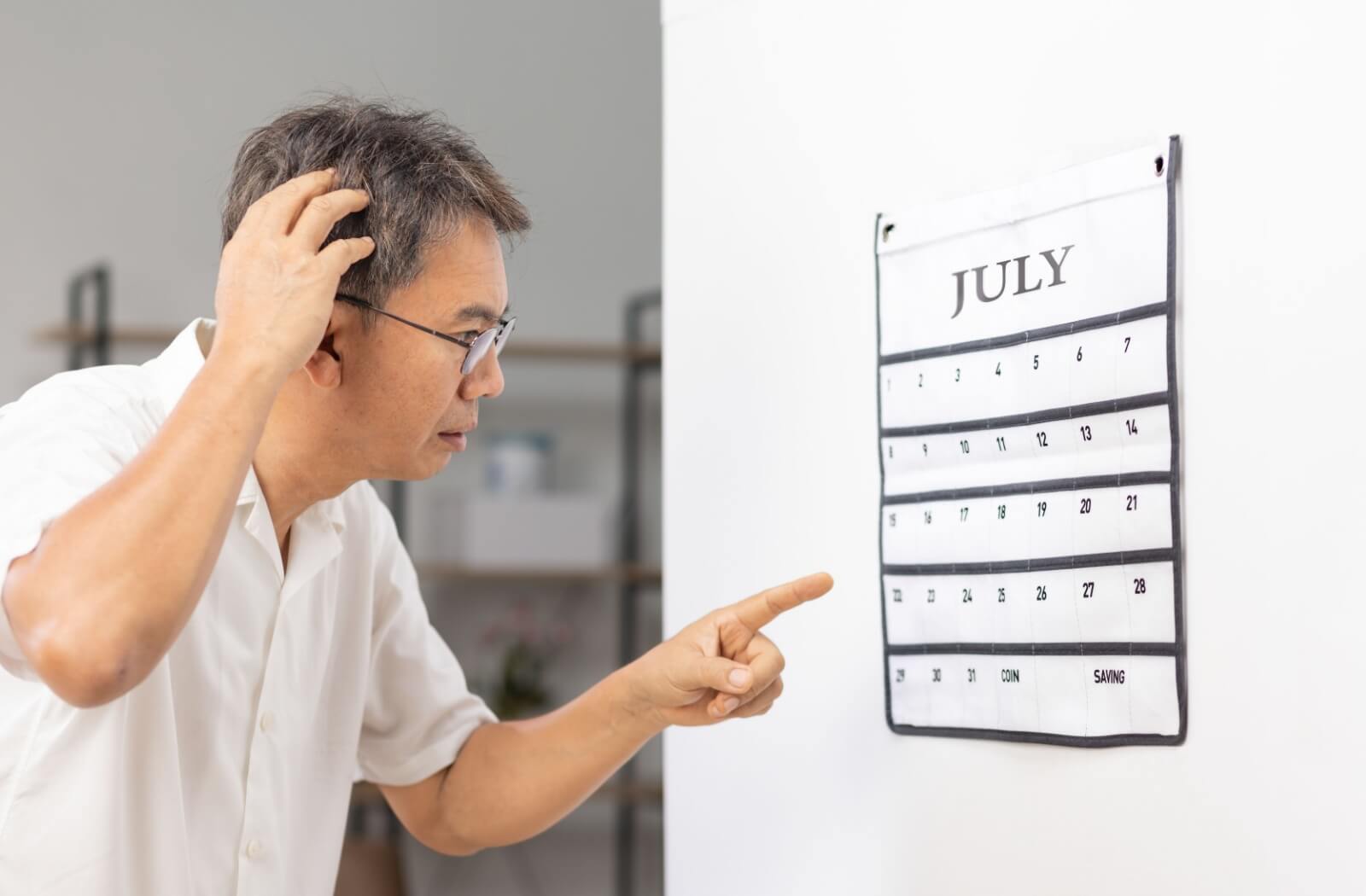 An older adult man, who is trying to remember an important date, pointing to a calendar on a wall.