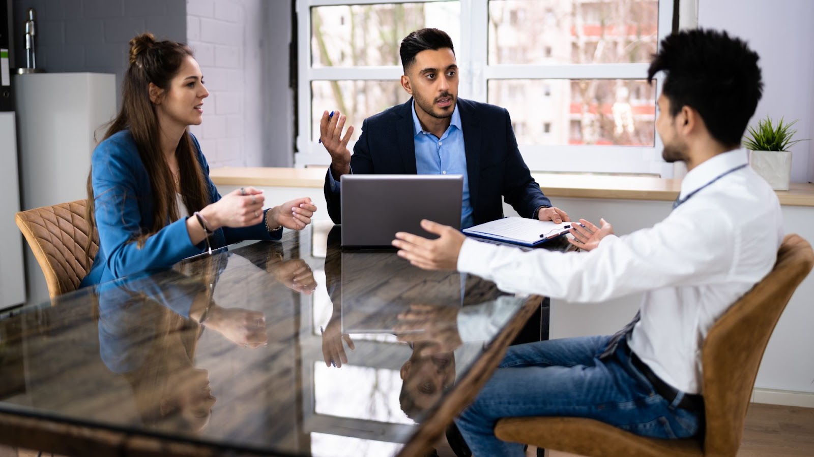 Three professionals engaged in a serious discussion around a reflective table in a modern office, with one person gesturing while holding a laptop.