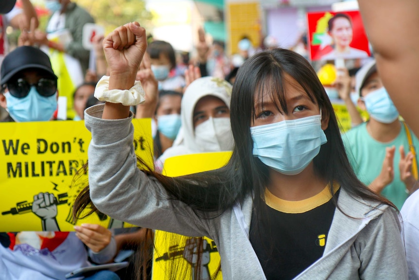 A woman with black hair and wearing a mask and jumper holds her fist in the air surrounded by a crowd of protesters.