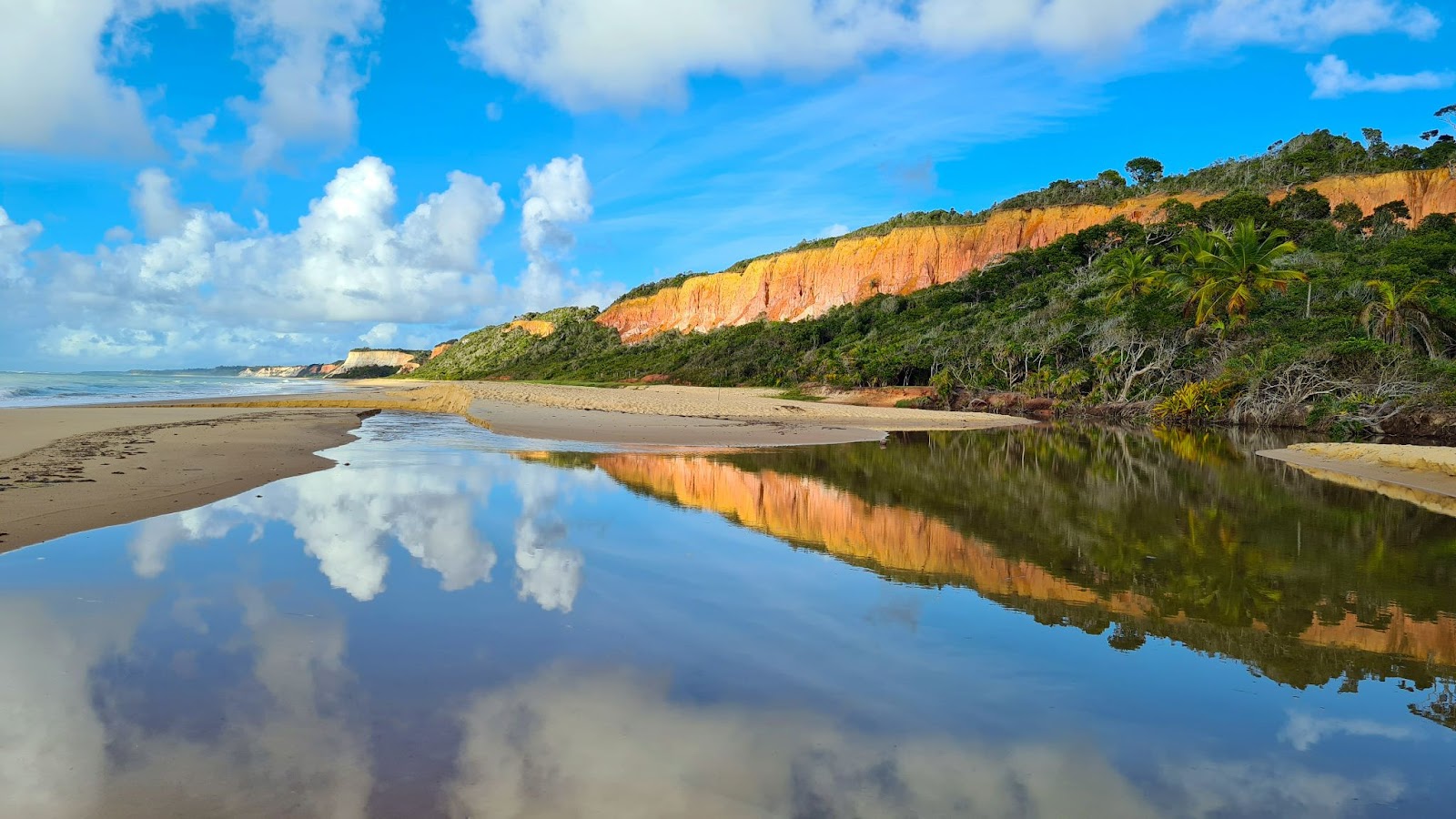 Piscina natural na areia da Praia da Pitinga refletindo o céu azul com nuvens brancos e a falésia coberta pela vegetação.