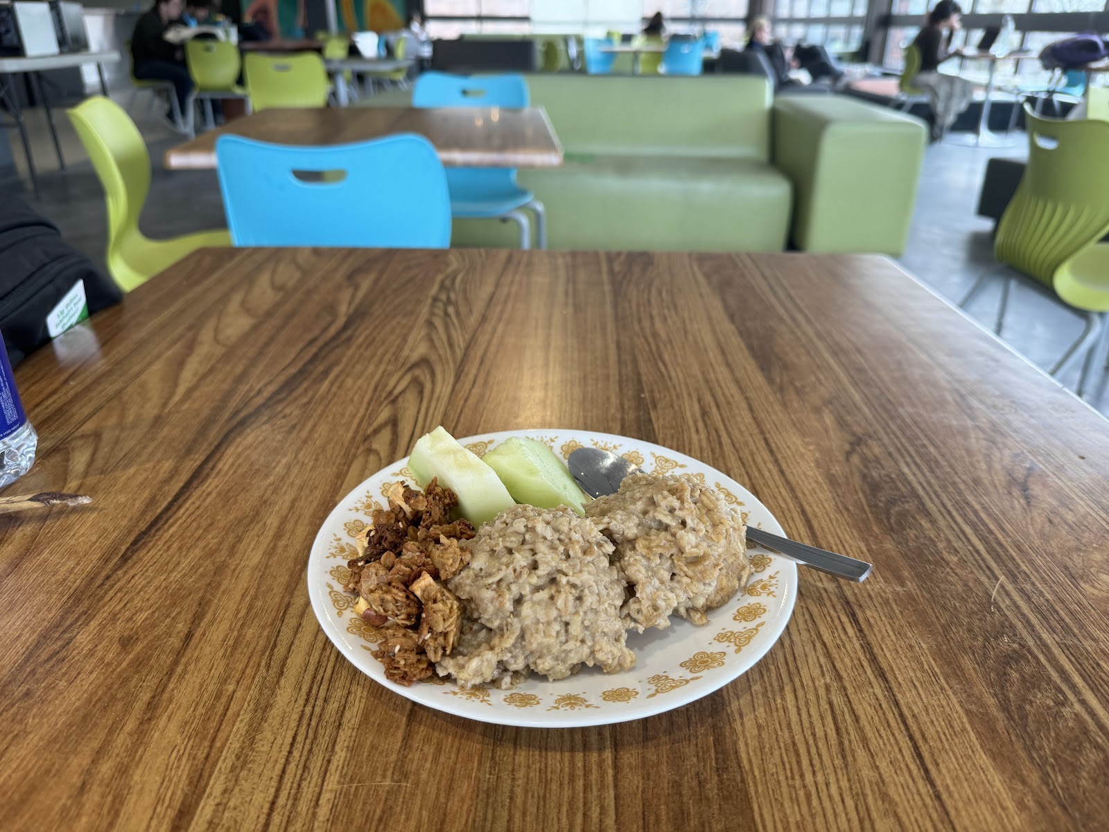 A plate of granola, oatmeal and honeydew melon on a wooden table. Blue and green chairs and sofas are visible in the background.