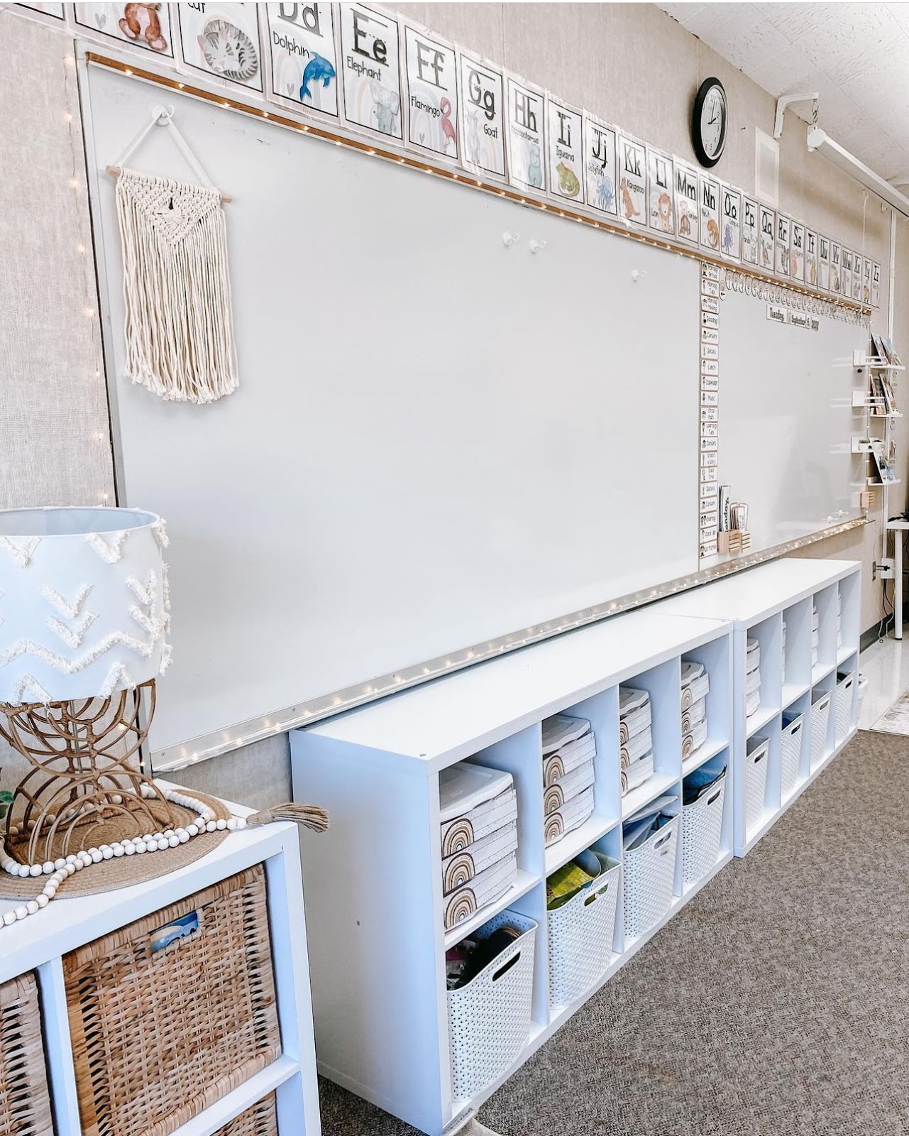 This image shows the front of a classroom with a boho rainbow classroom theme. There are soft, twinkle lights surrounding the bulletin board and lots of whites, browns, and other neutral colors. 